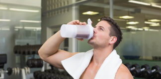 Suplementación con creatina para un mejor rendimiento físico y mental Shirtless male with white towel standing in dumbbell area of gym raising translucent shaker bottle. Athlete, fitness, strength, training, workout, recovery, gym