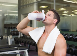 Suplementación con creatina para un mejor rendimiento físico y mental Shirtless male with white towel standing in dumbbell area of gym raising translucent shaker bottle. Athlete, fitness, strength, training, workout, recovery, gym