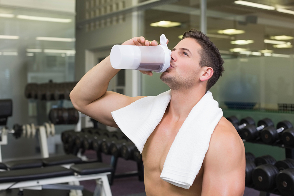 Shirtless male with white towel standing in dumbbell area of gym raising translucent shaker bottle. Athlete, fitness, strength, training, workout, recovery, gym